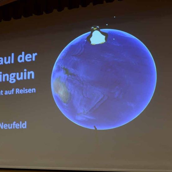 Eine Folie zeigt eine Erde mit einem Pinguin darauf, beschriftet mit 'Paul der Pinguin auf Reisen'. Die Erde ist blau und zeigt die Erde.