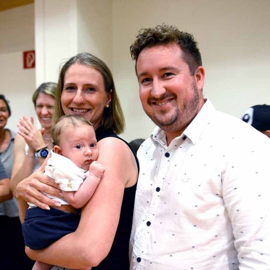 Eine lächelnde Frau hält ein Baby, während sie neben einem lächelnden Mann in einem weißen Hemd mit kleinen blauen Formen steht. Sie sind von anderen lächelnden Menschen in einem Raum umgeben.