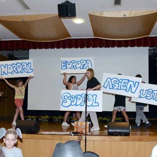Mehrere Kinder halten Schilder mit Namen von Kontinenten auf der Bühne hoch, mit Lautsprechern und einer Gitarre auf dem Bühnenboden.