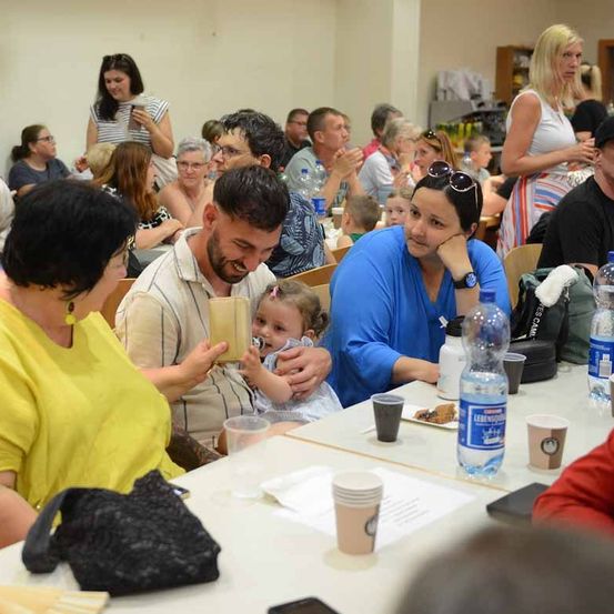 Eine Gruppe von Menschen sitzt an einem langen Tisch. Ein Mann hält ein Kind und schaut es an, während eine Frau die Hand des Kindes hält. Wasserflaschen stehen auf dem Tisch.