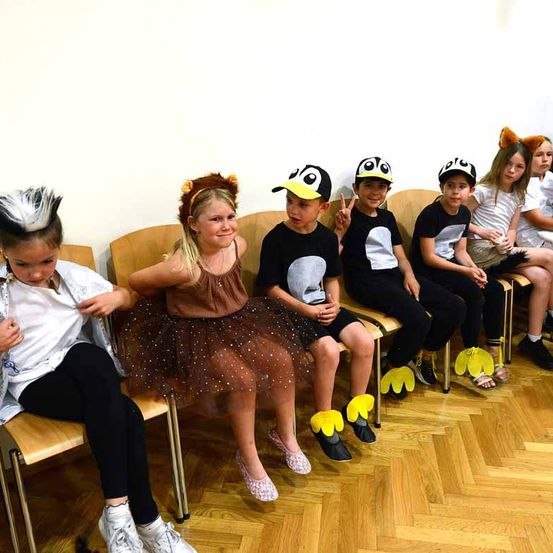 Mehrere Kinder sitzen in einer Reihe auf Stühlen und tragen verschiedene tierische Kostüme und Schuhe. Die Kinder lächeln, eines macht ein Friedenszeichen. Der Hintergrund ist eine schlichte weiße Wand.