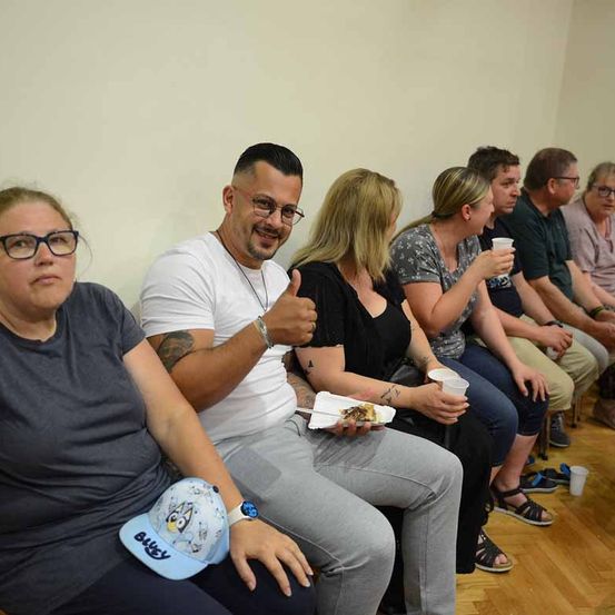 Eine Gruppe von Menschen sitzt auf einer Bank, einige mit Brille, einer gibt Daumen hoch. Zwei Frauen haben Tassen in den Händen. Eine trägt eine Halskette und hält Essen auf einem Teller.