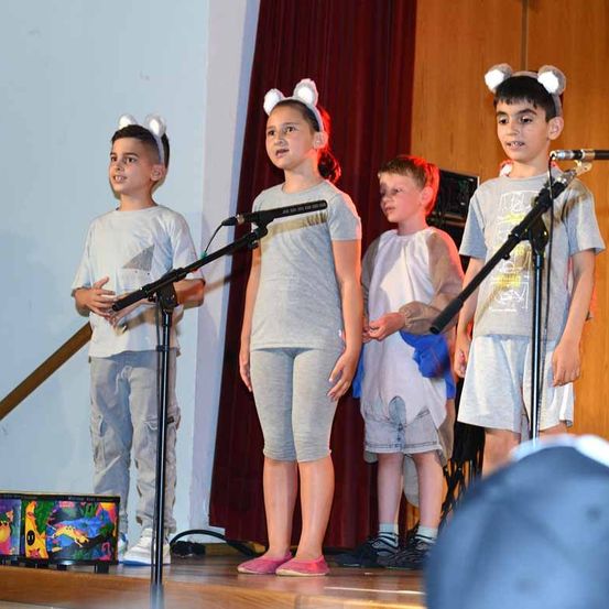 Vier Kinder, die Katzenohren tragen, stehen mit Mikrofonen auf der Bühne. Hinter ihnen befindet sich eine Holzwand mit einem roten Vorhang. Ein Kind hält eine bunte Trommel.