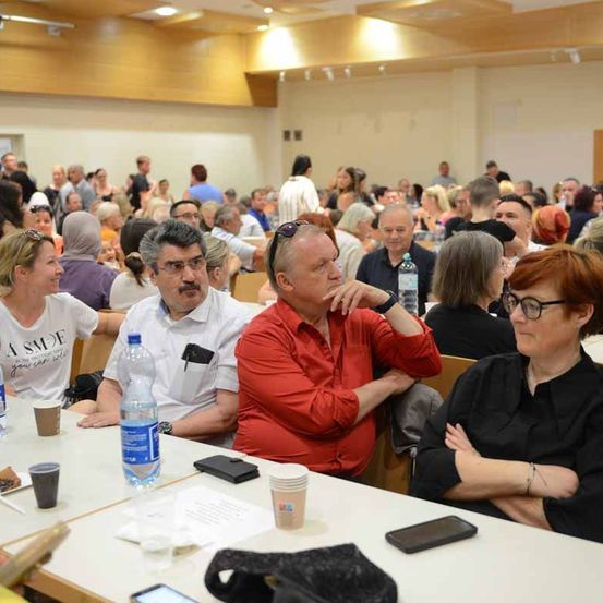 Ein Konferenzraum ist mit vielen Teilnehmern gefüllt, die an Tischen sitzen. Ein Mann in einem roten Hemd stützt sein Kinn auf seine Hand. In der Nähe hat eine Frau mit Brille die Arme verschränkt. Auf den Tischen befinden sich Becher, Flaschen und Telefone.