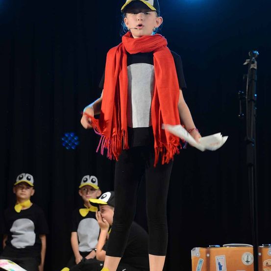 Ein junges Mädchen steht auf der Bühne und singt, während sie ein Papier in der Hand hält. Sie trägt einen roten Schal und schwarze Hosen. Hinter ihr sitzen drei Kinder auf dem Boden, die Pinguinkostüme tragen.