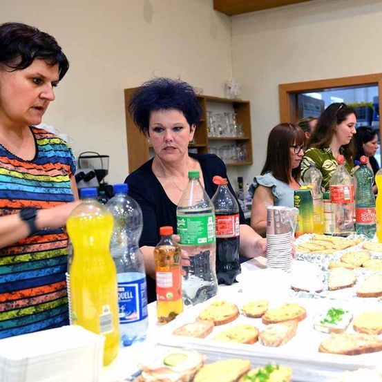 Mehrere Frauen bereiten Essen in einer Küche zu. Ein Tisch ist mit verschiedenen Lebensmitteln und Getränken beladen. Zwei Frauen stehen eng beieinander, während andere das Essen beobachten.