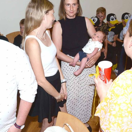 Eine Frau, die ein Baby hält, steht mit einer anderen Frau in einem Raum mit Kindern in Pinguinkostümen. Ein Mann in einem weißen Hemd steht links.