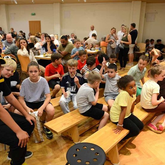 Eine große Gruppe von Kindern und Erwachsenen sitzt auf Holzbänken in einem Saal, einige essen, andere lauschen. Flaschen und andere Gegenstände stehen auf den Tischen. Ein Mann in einem gelben Shirt steht in der Nähe.
