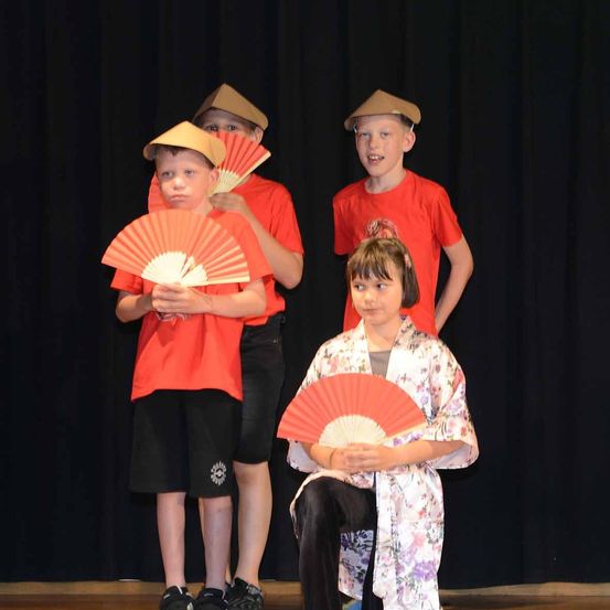 Vier Kinder auf der Bühne, zwei kniend, zwei stehend, tragen orangefarbene T-Shirts, Hüte und halten rote Fächer. Der Hintergrund ist schwarz.