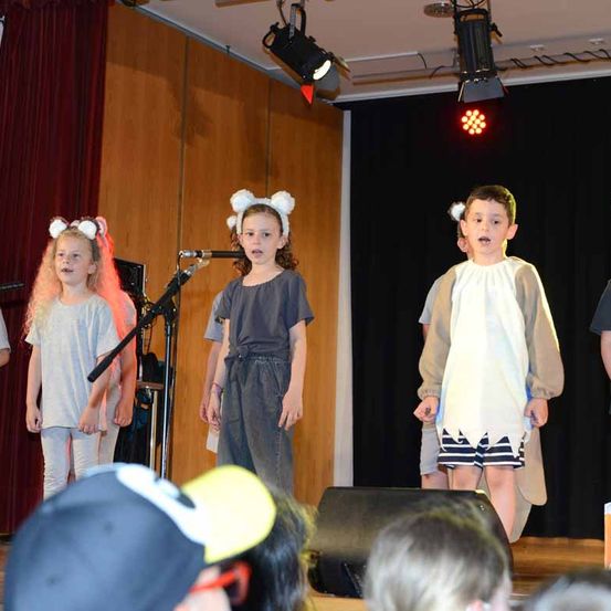 Drei Kinder stehen auf der Bühne und führen vor einem Publikum auf. Das Mädchen links trägt eine rosa Perücke und der Junge rechts ein weißes Gewand. Das Mädchen in der Mitte singt in ein Mikrofon.