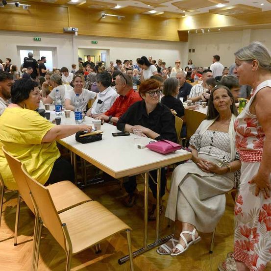 Ein voll besetzter Raum mit Holzböden und Stühlen. Die Leute sitzen an Tischen, und einige stehen. Eine Frau in einem Blumenkleid steht neben einer sitzenden Frau mit Brille und einer Handtasche.