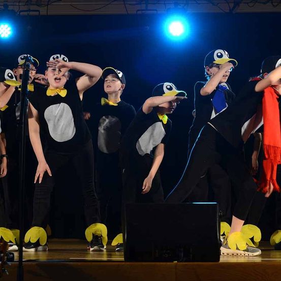 Eine Gruppe von Kindern, die als Pinguine verkleidet sind, tritt auf einer Bühne mit blauer Beleuchtung auf, einer singt in ein Mikrofon.