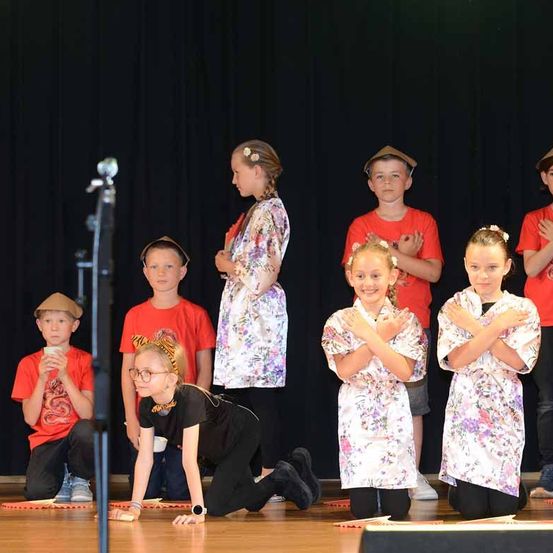Eine Gruppe von Kindern auf der Bühne, einige in roten T-Shirts und andere in weißen Blumenroben, führen ein Theaterstück auf. Ein Kind kniet auf dem Boden und andere klatschen.