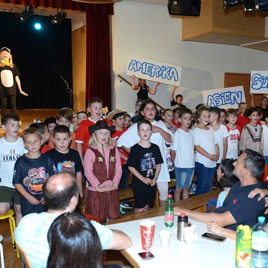 Eine Gruppe von Kindern versammelt sich in einem Raum. Sie halten Schilder hoch, auf denen 'Amerika' und 'Asiatisch' steht. Einige von ihnen singen. Vor ihnen sitzen zwei Erwachsene an einem Tisch mit Flaschen und einem Handy. Dahinter befindet sich eine Bühne mit einer Person, die ein Schild hält.