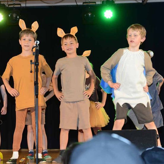 Drei Jungen stehen auf der Bühne mit Tierohren. Einer von ihnen trägt ein braunes Shirt und Shorts. Hinter ihnen führen andere eine Aufführung durch. Ein Mikrofon und Scheinwerfer sind vorhanden.