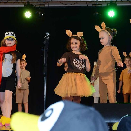 Kinder in Tierkostümen treten auf einer Bühne auf. Ein Mädchen in einem braunen Shirt mit Leopardenmuster, ein Mädchen in einem braunen Kleid und ein Mädchen in einem Pinguinkostüm sind auf der Bühne. Dahinter stehen zwei Jungen.