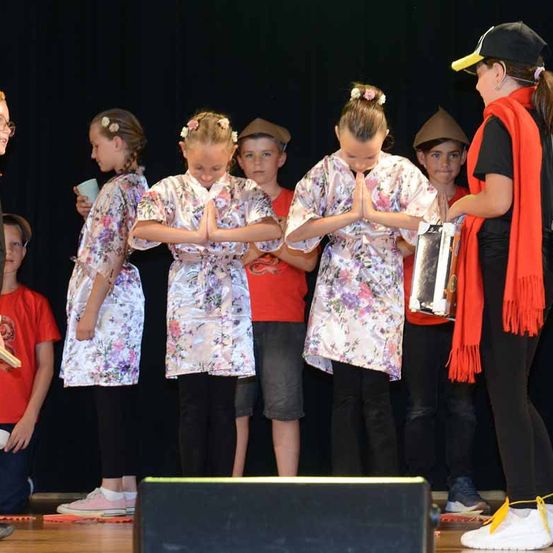 Mehrere Kinder in blumigen Kostümen stehen auf einer Bühne. Ein Mädchen hält einen Becher, ein anderes ein Buch. Eine erwachsene Frau steht neben ihnen und trägt einen roten Schal.