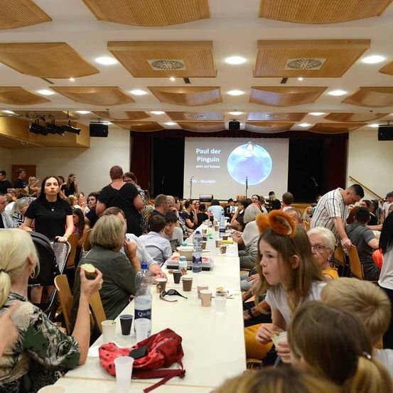 Ein Konferenzsaal mit einer Projektionsleinwand, die Paul der Pingu zeigt. Die Teilnehmer sitzen um Tische herum, einige essen. Der Saal hat Holzdecken und eine Bühne mit einem Projektor.
