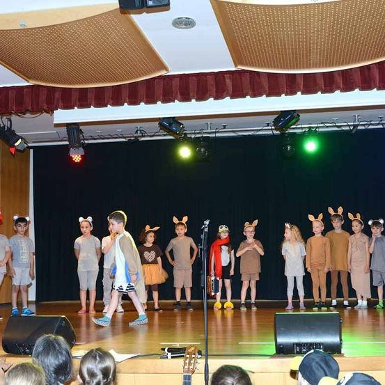 Kinder in Tierkostümen führen eine Aufführung auf der Bühne durch, begleitet von einer Lehrerin. Sie stehen in einer Reihe, beleuchtet von Bühnenlampen.