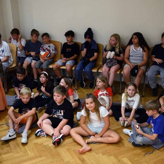 Kinder sitzen in einem Kreis auf dem Boden in einem Raum. Einige schauen auf etwas, während andere wegschauen. Einige tragen Turnschuhe.