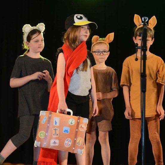 Vier Kinder auf der Bühne, in Kostümen gekleidet. Das Mädchen in der Mitte hält einen Koffer mit Aufklebern von verschiedenen Orten. Der Junge rechts trägt ein Mikrofon. Das Mädchen links trägt einen Hasenohr-Kopfschmuck.
