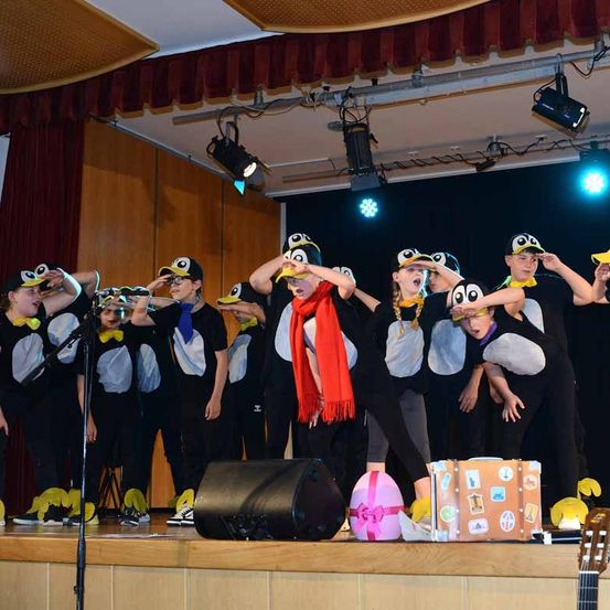 Eine Gruppe von Kindern in Pinguinkostümen tritt auf der Bühne auf, mit verschiedenen Requisiten wie einem Koffer und einem rosa Ball.