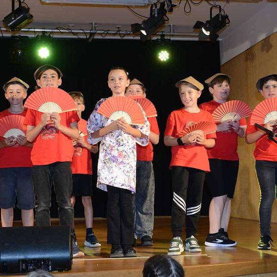 Eine Gruppe von Kindern auf der Bühne hält rote Fächer. Sie tragen rote T-Shirts und Hüte, zwei halten Mikrofone. Lichter und ein Lautsprecher stehen hinter ihnen.