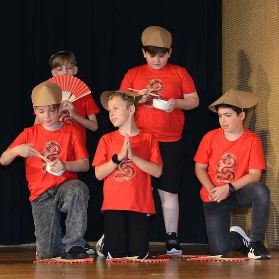 Fünf Jungen in roten Hemden und Hüten treten auf einer Bühne auf, einer mit einer Schüssel und Chopsticks, ein anderer hält einen Fächer. Sie scheinen an einer kulturellen Veranstaltung teilzunehmen.