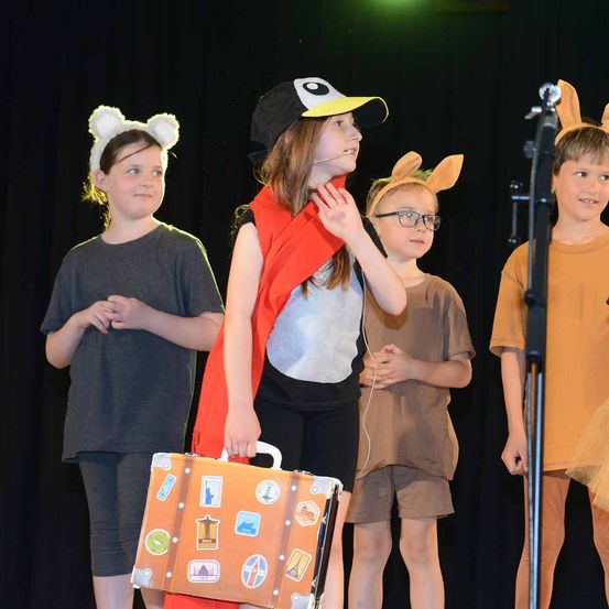 Vier Kinder auf der Bühne in Kostümen, eines mit einem Koffer, zwei mit Tierohren und eines mit einem Mikrofon.