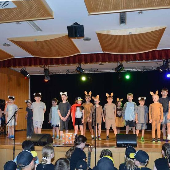 Kinder in Tierkostümen spielen auf einer Bühne mit Scheinwerfern und Lautsprechern. Das Publikum beobachtet von unten.