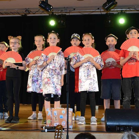 Eine Gruppe von Kindern in verschiedenen Kostümen steht auf der Bühne und hält rote Fächer. Einige tragen Hüte, und es gibt Scheinwerfer und einen Lautsprecher im Hintergrund.