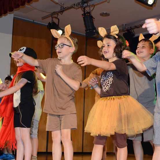 Kinder in Tierkostümen treten auf einer Bühne auf, mit Scheinwerfern im Hintergrund.