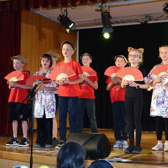 Eine Gruppe von Kindern in roten Hemden und Mützen tritt mit roten Fächern auf der Bühne auf. Ein Mikrofon steht vor ihnen. Scheinwerfer sind an der Decke montiert.