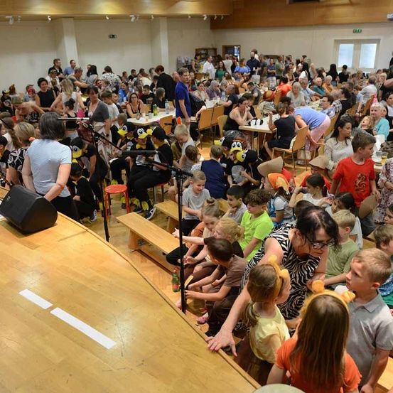 Ein überfüllter Raum mit Holzbänken und Tischen, einer Person bei einem Mikrofon und sitzenden und stehenden Kindern und Erwachsenen. Einige tragen Kostüme.