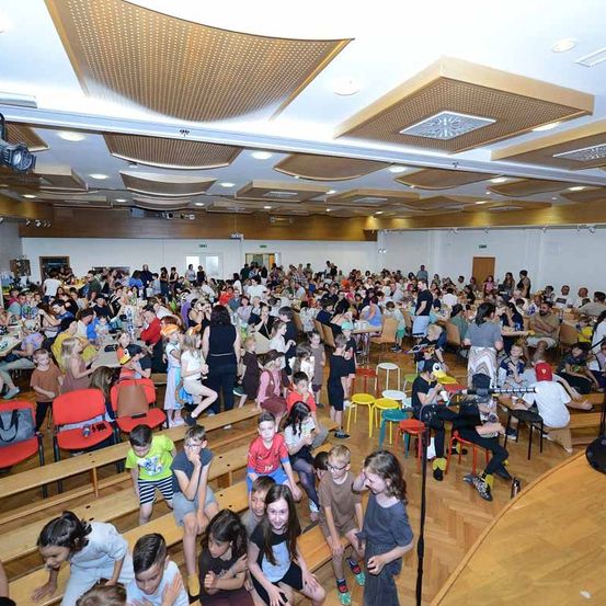 Eine große Gruppe von Menschen in einem Konferenzsaal. Einige sitzen, andere stehen und eine Person sitzt an einem Tisch. Der Raum hat Holzböden und Stühle.