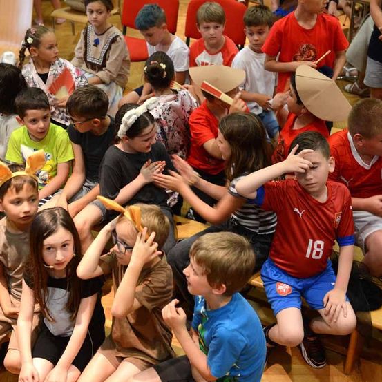 Eine Gruppe von Kindern sitzt auf Bänken und trägt Hasenohren und Papierhüte. Ein Junge in einem roten Trikot mit der Nummer 18 hat die Hand auf der Stirn.