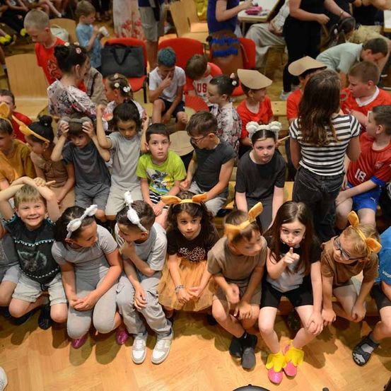 Eine Gruppe von Kindern in Kostümen mit Hasenohren sitzt auf dem Boden, möglicherweise bei einer Aufführung. Einige haben die Hände auf dem Kopf. Dahinter sitzen andere auf Stühlen. Im Hintergrund sitzen weitere Kinder auf Bänken.