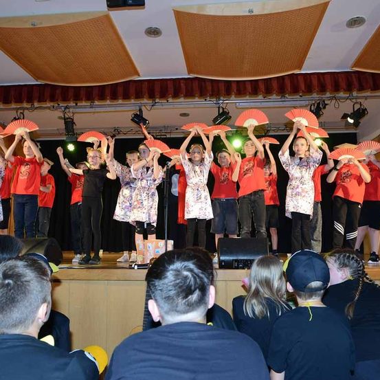 Eine Gruppe von Kindern in roten Hemden und schwarzen Hosen führt mit roten Fächern auf der Bühne auf. Die Bühne hat einen Holzboden und einen schwarzen Vorhang. Scheinwerfer beleuchten die Darsteller. Das Publikum sieht von vorne zu.