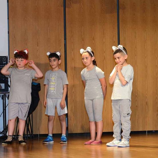 Vier Kinder, die Tierohren tragen, stehen auf der Bühne, wahrscheinlich in einem Schulstück. Sie tragen graue T-Shirts und Shorts. Der Junge auf der linken Seite hat rote Haare. Hinter ihnen ist eine Holzwand.