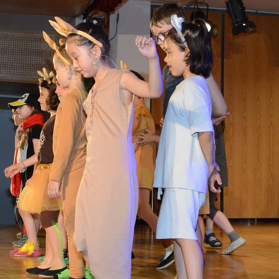 Mehrere Kinder führen auf einer Bühne auf und tragen Kostüme. Ein Mädchen vorn trägt ein braunes Kleid und eine Kopfbedeckung mit Hasenöhrchen. Dahinter tragen andere Kinder verschiedene Kostüme. Die Bühne hat einen Holzboden.