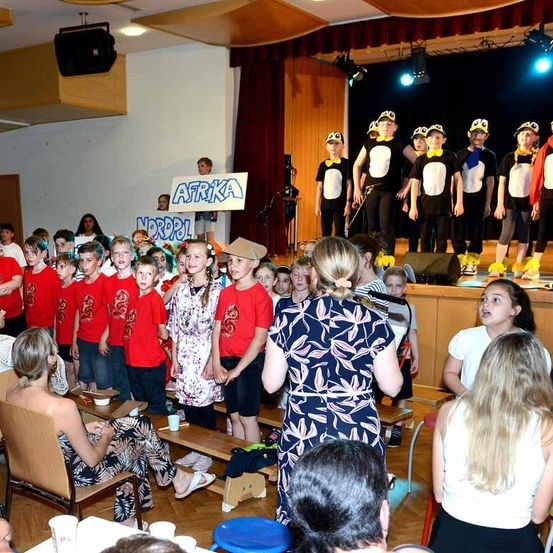 Eine Gruppe von Kindern in roten Hemden tritt auf der Bühne auf, vor ihnen eine Frau. Sie halten ein Schild mit der Aufschrift 'Afrika'. Auf der Bühne stehen auch andere Kinder in Pinguinkostümen. Daneben beobachten andere Menschen. Es gibt Holzbänke, Tische und Stühle im Bereich.