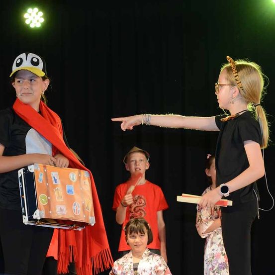 Ein Mädchen mit einem Koffer und einer Pinguinmütze steht auf der Bühne. Ein weiteres Mädchen mit einem Tigerkopfband zeigt zur Seite. Ein Junge in einem roten Shirt und ein Mädchen in einem Blumenkleid stehen ebenfalls auf der Bühne.