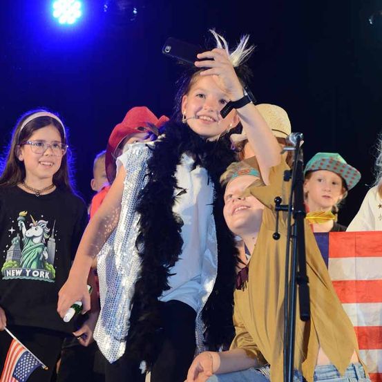 Eine Gruppe von Kindern auf der Bühne, eines der Kinder macht ein Selfie. Sie sind in Kostümen gekleidet.