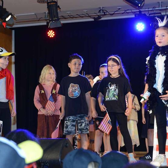 Eine Gruppe von Kindern auf der Bühne, einige in Kostümen und mit Flaggen, mit Bühnenlicht im Hintergrund.