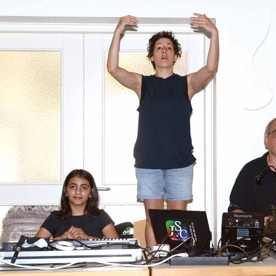 Eine Frau steht mit erhobenen Armen in einem Raum mit einer Glastür, neben einem sitzenden Mädchen und einem Mann mit einem Mikrofon. Der Tisch vor ihnen ist mit Laptops, Kabeln und anderen Geräten übersät.