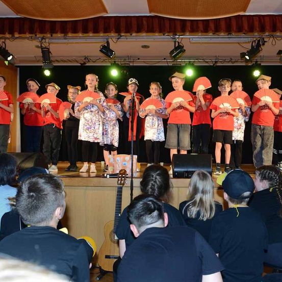 Eine Gruppe von Kindern in roten Hemden und Mützen tritt mit Fächern auf der Bühne auf, begleitet von einer Person mit Mikrofon. Ein Publikum sitzt davor, und eine Gitarre ist auf der Bühne platziert. Scheinwerfer und Lautsprecher sind um die Bühne herum positioniert.