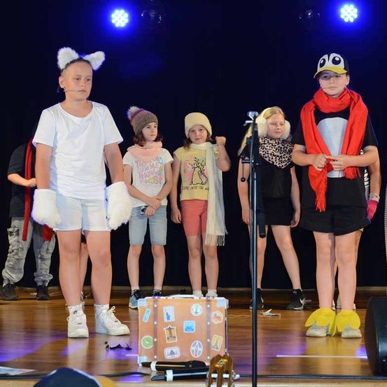 Auf einer Bühne stehen mehrere Kinder um einen Koffer mit Aufklebern und tragen Kostüme. Einer hat eine Katzenohren-Kopfbedeckung, ein anderer eine Pinguin-Mütze. Der Hintergrund ist dunkel mit blauen Lichtern.