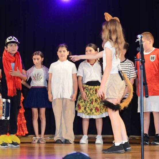 Eine Gruppe von Kindern steht auf der Bühne, einige in Kostümen. Ein Mädchen in einem weißen Shirt und beige Shorts hält ein Requisit. Die Bühne ist schwach beleuchtet, und der Hintergrund ist dunkel.