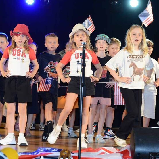 Eine Gruppe von Kindern steht mit amerikanischen Flaggen auf der Bühne, trägt USA- und Arizona-T-Shirts. Ein Mädchen steht vor einem Mikrofon, während andere Flaggen halten. Ein Lautsprecher steht rechts.