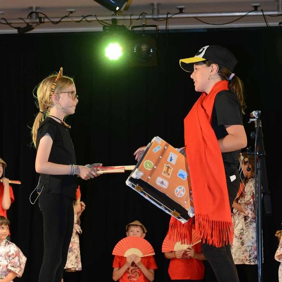 Ein Mädchen mit Brille überreicht einer anderen Frau in einem schwarzen Hut auf der Bühne ein Buch, beide halten Requisiten. Hinter ihnen beobachten mehrere Kinder in Kostümen, einige halten Fächer. Scheinwerfer und schwarze Vorhänge im Hintergrund.